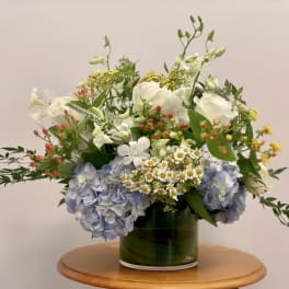 Low glass vase of white roses, blue hydrangeas, and small white daisies with berries on a wooden table