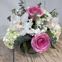 Low arrangement of pink roses, white hydrangeas, stock, and orchids in a clear glass vase