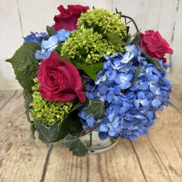 Low round arrangement of hot pink roses, green blooms, and blue hydrangeas in a clear glass vase