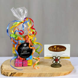 Bag of colorful wrapped milk chocolates with curling ribbons on a wooden board beside a florist business card.