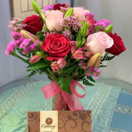 Bouquet of red and pink roses with lilies in a glass vase, with a candy box below