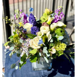 Mixed arrangement of white roses and calla lilies with purple and blue blooms in a square glass vase