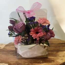 Pink and purple flower arrangement in a clear gift box with a ribbon bow