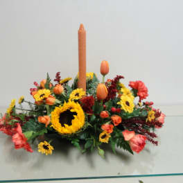 Low floral centerpiece with a tall orange candle and mixed orange, yellow, and red blooms