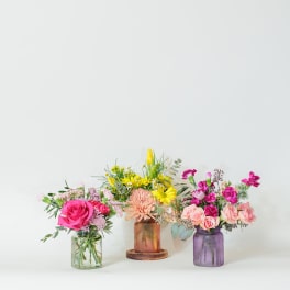 Three small mixed flower arrangements in colorful glass jar vases on a light background.