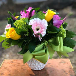 Bouquet of yellow roses, pink gerbera daisies, and purple flowers in a white vase