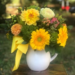 Yellow and peach flowers arranged in a white pitcher vase with a yellow ribbon