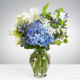 Arrangement of white lilies, blue hydrangeas, and purple flowers in a clear glass vase