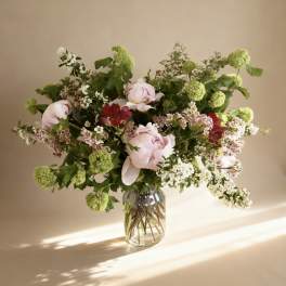 Loose arrangement of pale pink peonies, red blooms, and green flower clusters in a clear glass vase.