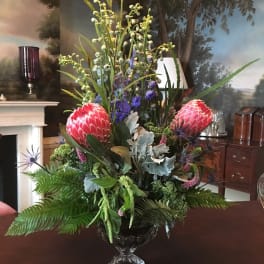 Tall tropical floral arrangement in a clear glass vase with pink protea and blue accents