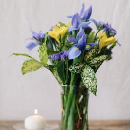 Blue and yellow flowers arranged in a clear glass vase beside a small candle