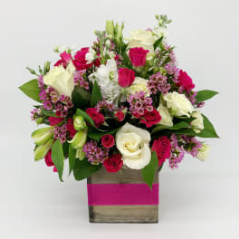 Compact arrangement of white and hot pink roses with small purple flowers in a wood cube vase wrapped with a pink band