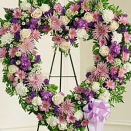 Heart-shaped standing wreath of pink, purple, and white flowers on a metal easel with a lavender bow.