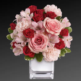 Compact arrangement of red and pink roses, alstroemeria, and carnations in a clear glass cube vase.