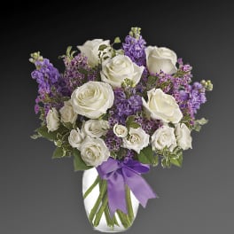 White roses and purple flowers arranged in a glass vase with a purple ribbon.