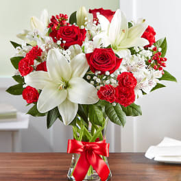Red roses, white lilies, and red carnations in a clear glass vase with a red ribbon bow