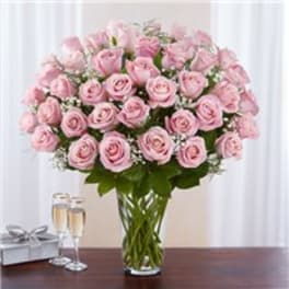 Large arrangement of pink roses with white filler flowers in a tall clear glass vase beside champagne flutes and a gift box