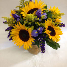 Arrangement of bright yellow sunflowers with purple irises in a glass vase wrapped in burlap.