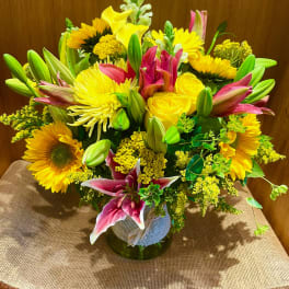 Mixed bouquet with yellow sunflowers, roses, lilies, and pink blooms in a glass vase