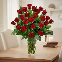 Tall bouquet of long-stemmed red roses in a clear glass vase on a dining table.