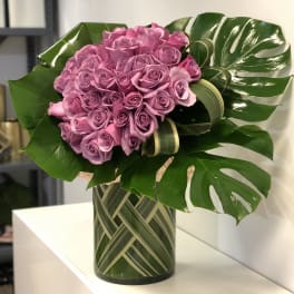 Tall modern arrangement of clustered lavender roses with large glossy leaves in a glass vase