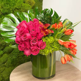 Bright pink roses, orange tulips, and green hydrangeas arranged with large tropical leaves in a clear glass cylinder vase