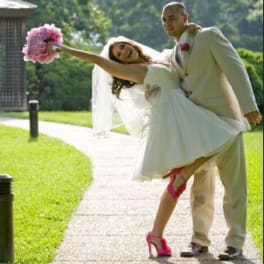 Bride being carried by groom, holding a pink bouquet outdoors