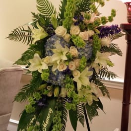 Tall standing spray of white lilies, roses, blue hydrangeas, and green bells of Ireland on an easel
