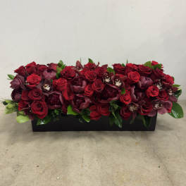 Low rectangular arrangement of dark red roses, peonies, and orchids in a black container
