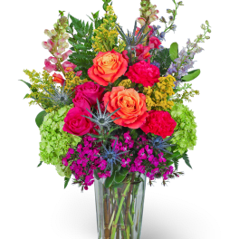 Tall arrangement of bright orange roses, hot pink blooms, and green hydrangeas in a clear glass vase.