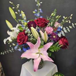 Mixed bouquet with pink lily, red roses, and blue accents in a glass vase