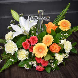 Low arrangement of white lily, peach gerberas, orange and pink roses with a clear glass cross in the center.