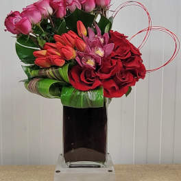 Pink and red floral arrangement in a tall glass vase with heart-shaped wire accents