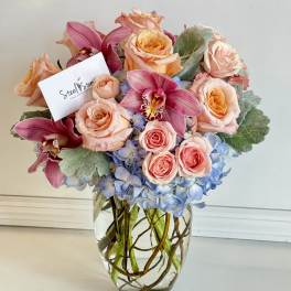 Pink roses and orchids arranged in a clear glass vase with blue hydrangeas