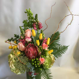 Tall mixed bouquet in a glass vase with pink roses, yellow tulips, and a protea