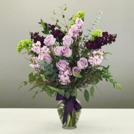Lavender roses and pink flowers in a glass vase with a purple ribbon