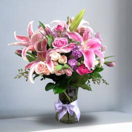 Pink lilies, roses, and purple flowers in a clear glass vase with a lavender ribbon.
