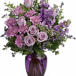 Purple and lavender roses, mums, and stock arranged in a purple glass vase.