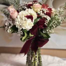 Tall vase arrangement of red and pink roses with white hydrangeas and baby's breath, tied with a burgundy bow.