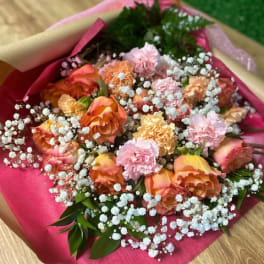 Hand-tied bouquet of peach-pink roses and carnations with white filler flowers in pink paper wrap