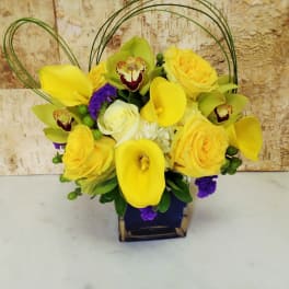 Low arrangement of yellow calla lilies, roses, and green orchids in a blue cube vase