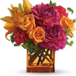Bright arrangement of orange lilies and roses with fuchsia dahlias in an orange glass cube vase