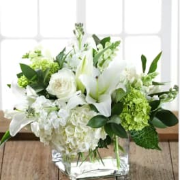 White lilies and roses arranged in a clear square vase with green accents