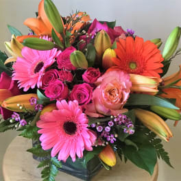 Bright mixed bouquet with pink gerberas, roses, and orange lilies in a glass vase.