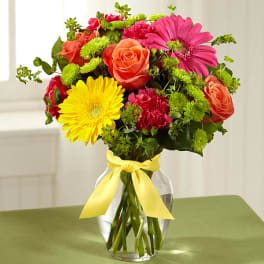 Mixed bouquet of roses, gerbera daisies, and carnations in a glass vase with a yellow ribbon.