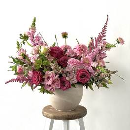 Pink mixed flower arrangement in a beige vase on a wooden stool