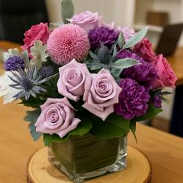 Compact arrangement of pink and purple flowers in a square glass vase on a wood slice.