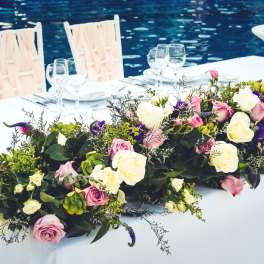 Long floral centerpiece with pink, white, and purple roses on a table by water