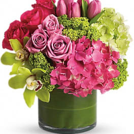 Compact pink and green arrangement with roses, tulips, orchids, and hydrangeas in a leaf-wrapped glass vase