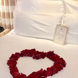 Red rose petals arranged in a heart on a bed with a "Will you marry me?" sign
